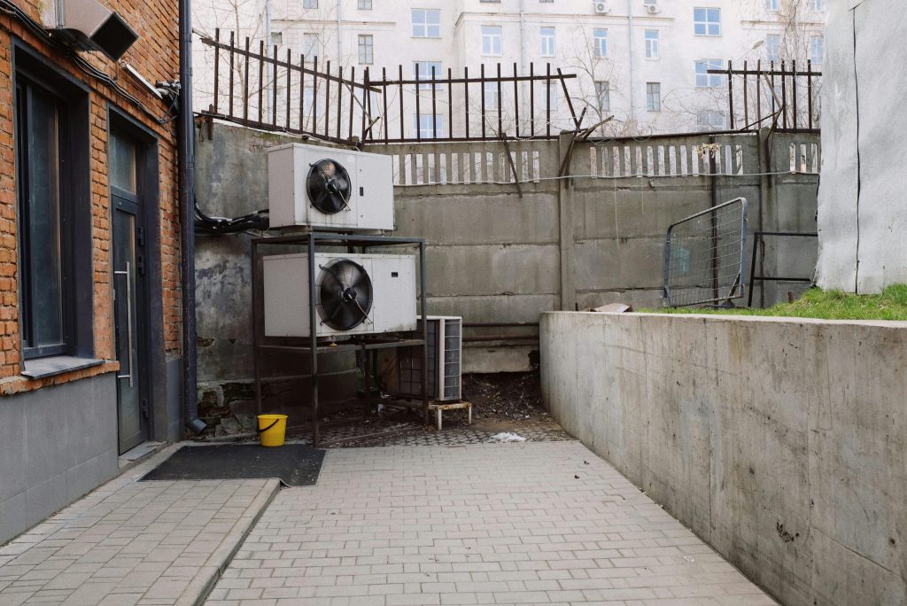 Air conditioning system located outside concrete shabby fence with metal barrier near industrial building from bricks and glass door